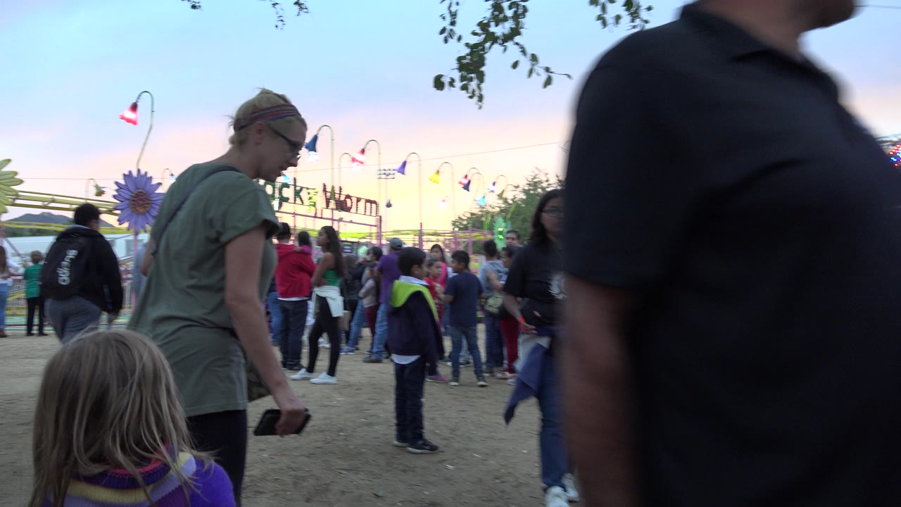 A dark-shirted passerby crosses close to the lens as a woman, child, and Wacky Worm ride crowd remain visible at the San Jose del Cabo fairground.