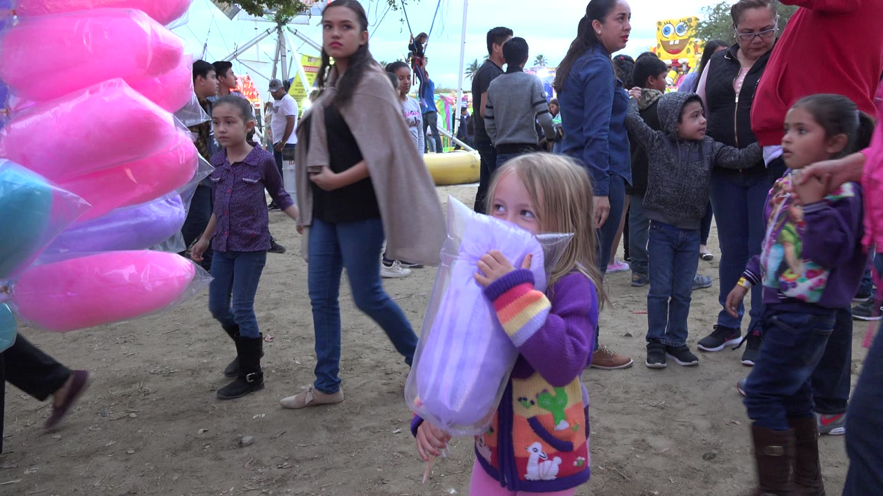 The girl stands closer to center in San Jose Del Cabo, Mexico with pale purple cotton candy in her arms, while children and adults gather around ride structures in the background.