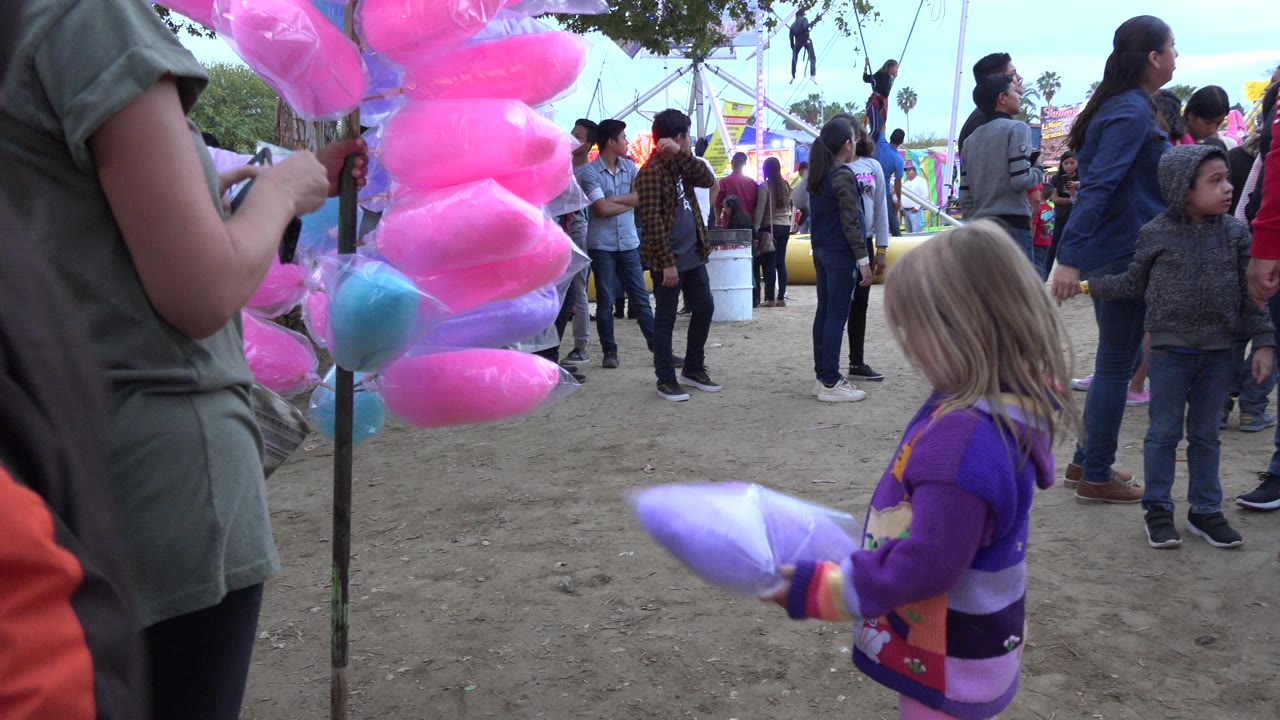 In San Jose del Cabo, Mexico, the girl holds the cotton candy lower in front of her as adults and children stand near colorful fair ride structures in the background.