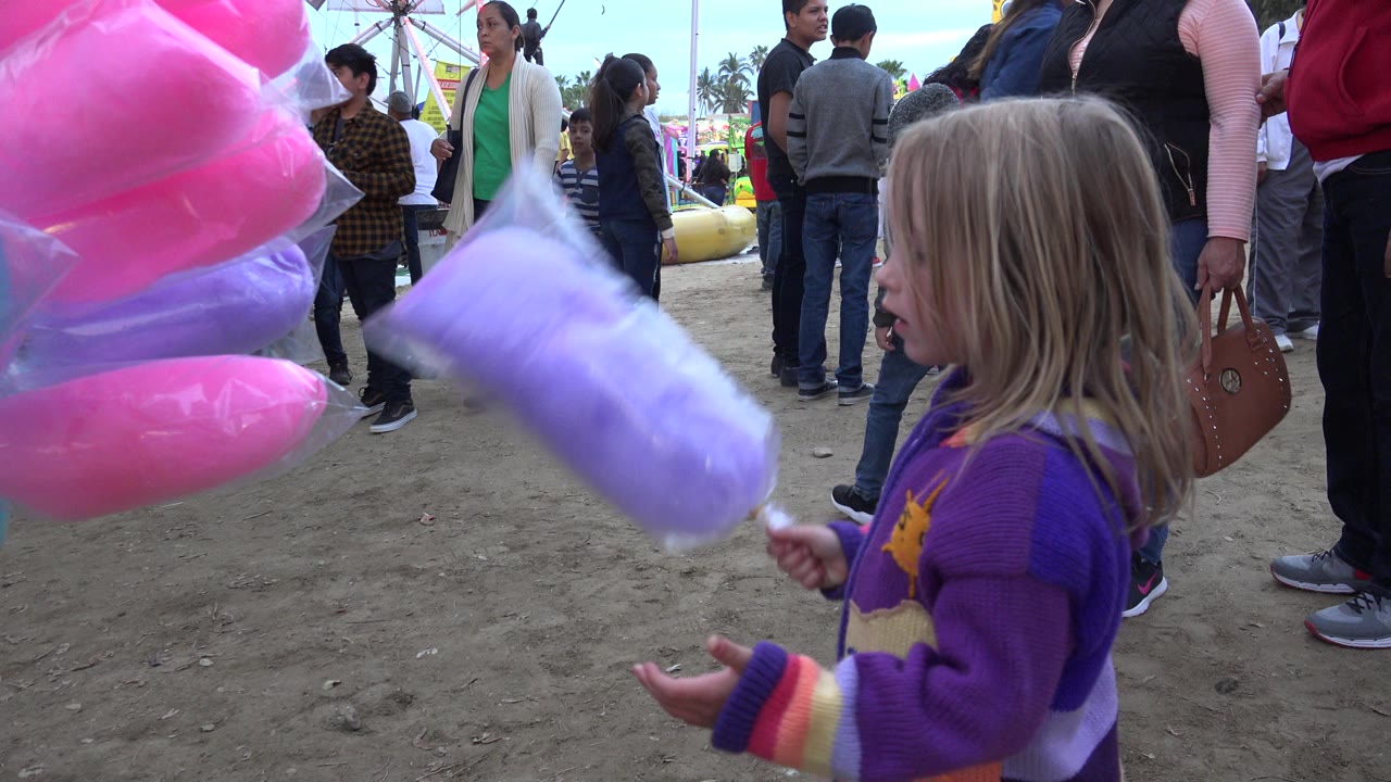Young girl holding purple cotton candy beside pink cotton candy at a crowded fair.