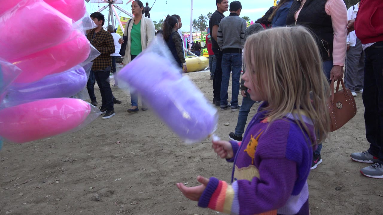 The girl remains in the foreground with purple cotton candy raised outward as adults and children stand across the fairground in San Jose del Cabo, Mexico.