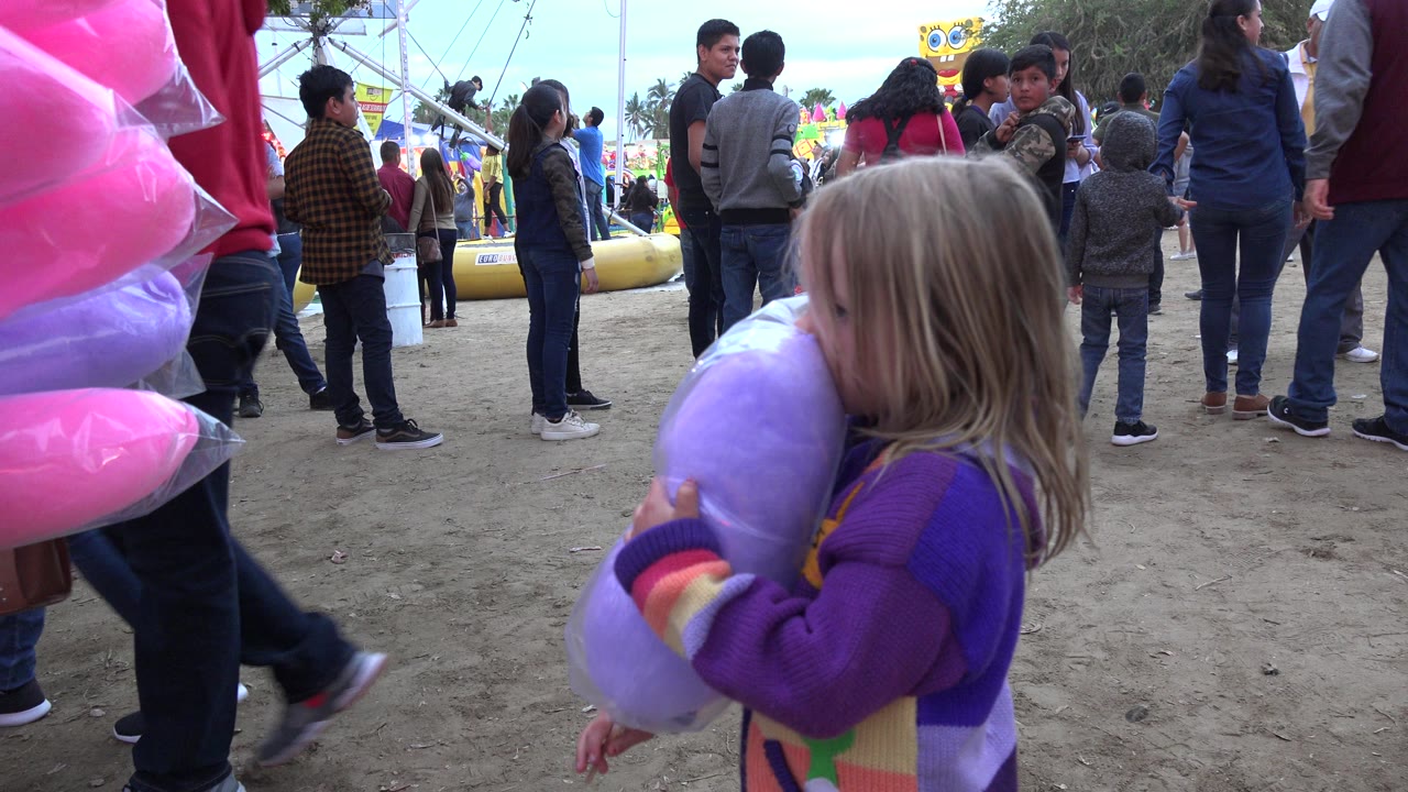 The girl keeps the purple cotton candy close while standing beside bright pink cotton candy bags, with a cluster of people and fair equipment in the San Jose Del Cabo background.