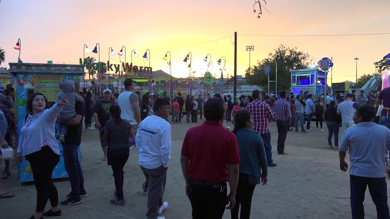 Families and visitors stand and walk near carnival booths in San Jose del Cabo, with a child on an adult's shoulders and the Wacky Worm sign lit against the evening sky.