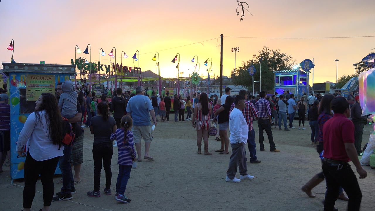 A dense group of pedestrians in San Jose del Cabo stands and walks near a Wacky Worm ride sign while the sky glows yellow and orange at dusk.