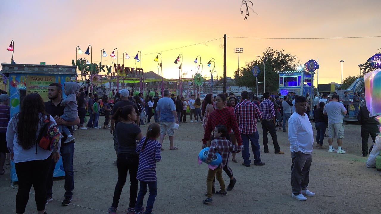 Crowd at an amusement park walkway at sunset with children and ride lights visible.