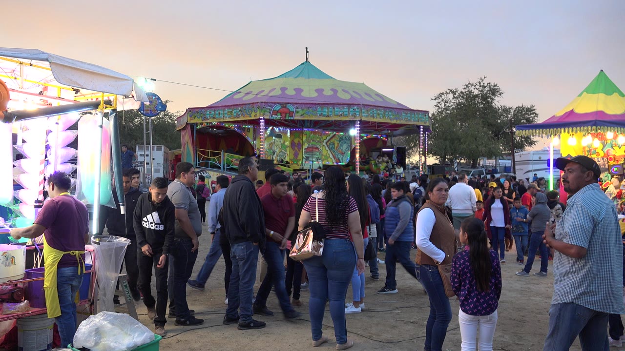 Crowded San Jose del Cabo fairground walkway at dusk with a bright booth on the left, colorful carnival tents in back, and families and teenagers facing different directions.