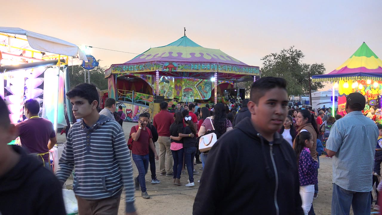 People and children gather at a San Jose del Cabo fairground, with a bright booth on the left, a painted pavilion in back, and a young man close to the camera in front.