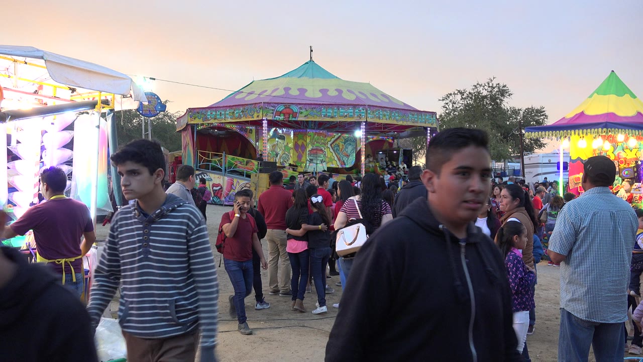 Two young men pass near the foreground at the San Jose del Cabo fair, while families stand between a bright booth at left and a colorful pavilion in the background.