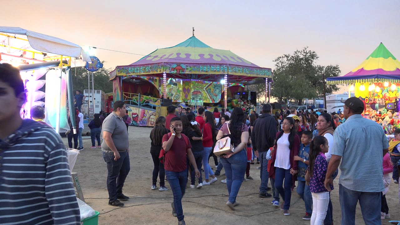 Families walk past fair booths and a colorful pavilion at dusk.