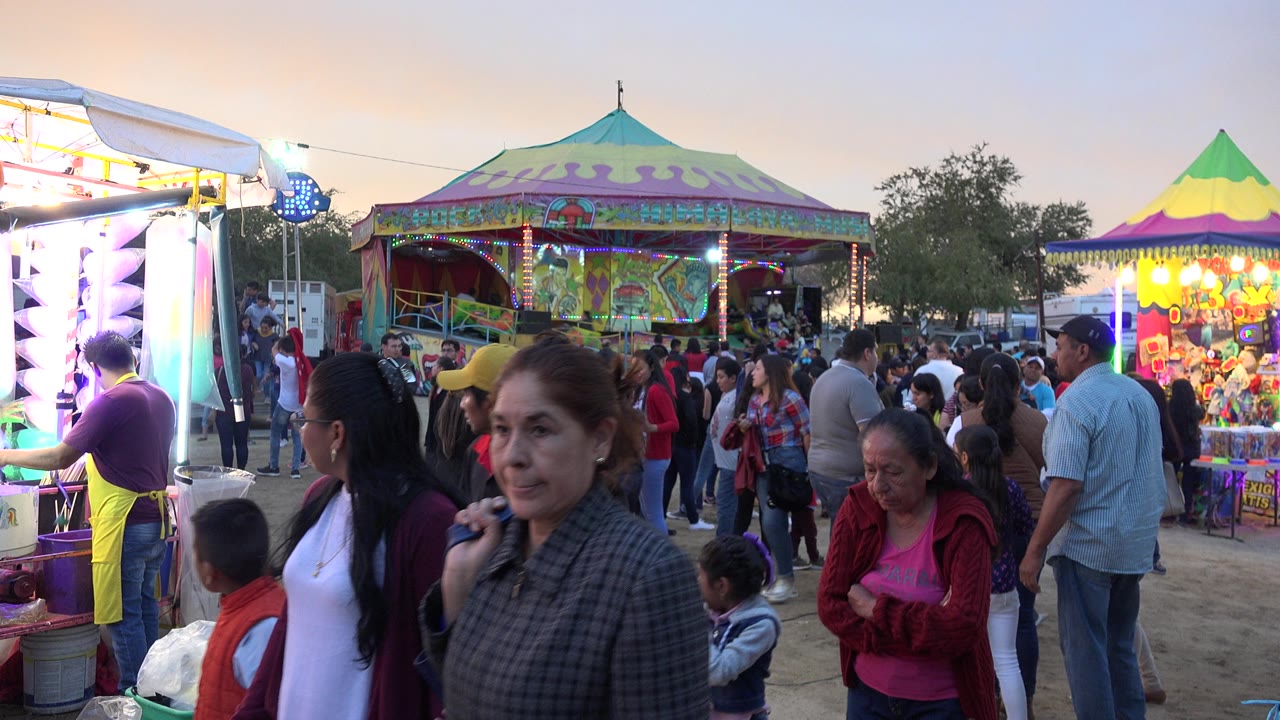 Crowded funfair walkway with adults, children, bright booths, and a covered ride.