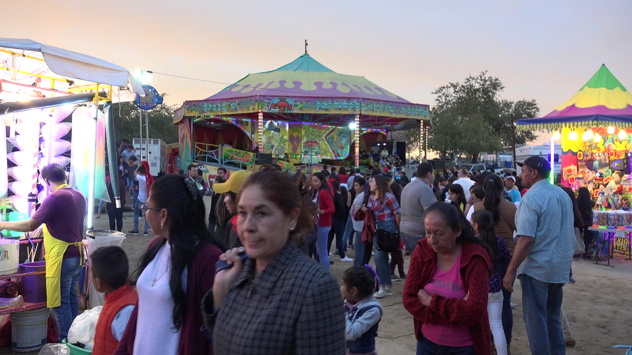 A woman passes near the foreground as families gather below bright funfair lights and a patterned ride canopy in San Jose del Cabo.