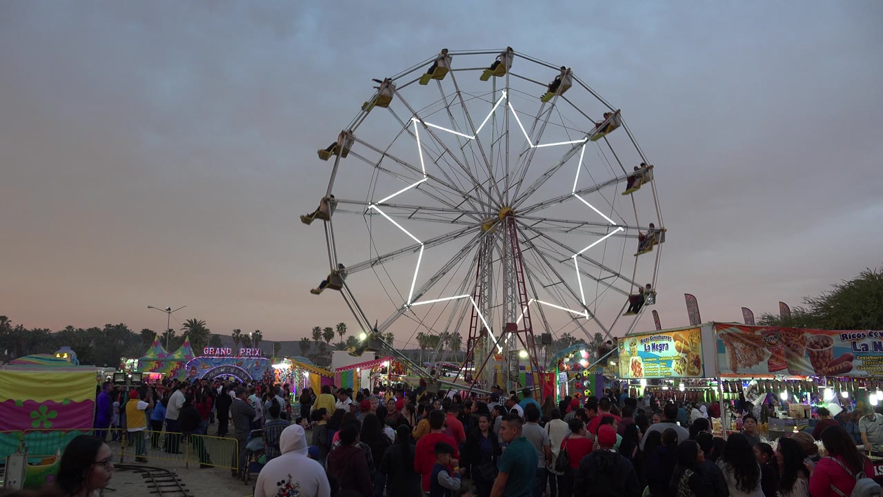Crowded fairground with a lit Ferris wheel under a cloudy evening sky.
