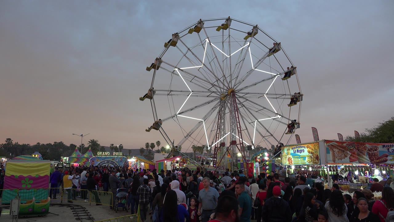 The Ferris wheel remains centered above the San Jose del Cabo fairground as the foreground crowd fills the walkway below colorful ride and food stalls.