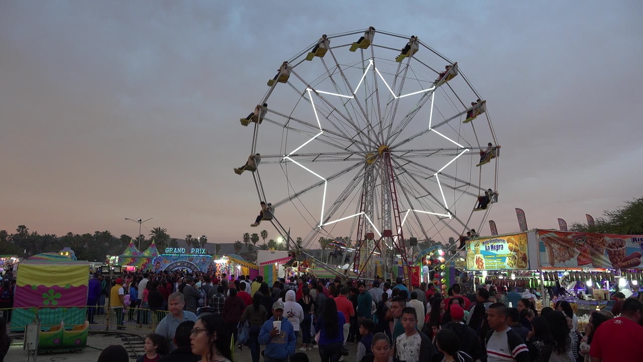 A crowded San Jose del Cabo fairground scene shows a large Ferris wheel with white lights above busy walkways and colorful stalls at dusk.