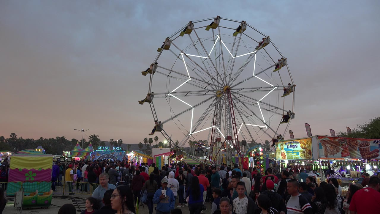 The San Jose del Cabo crowd fills the foreground while the lit Ferris wheel and bright booth signs sit behind them under a dim evening sky.