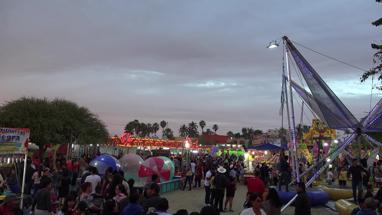 Families and children fill the San Jose del Cabo fairground walkway as neon booth lights glow behind them and the tall ride frame stands at the right.
