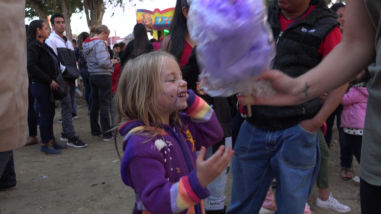 Young child in a purple sweater looks up near a purple cotton candy bag in a crowded outdoor area.