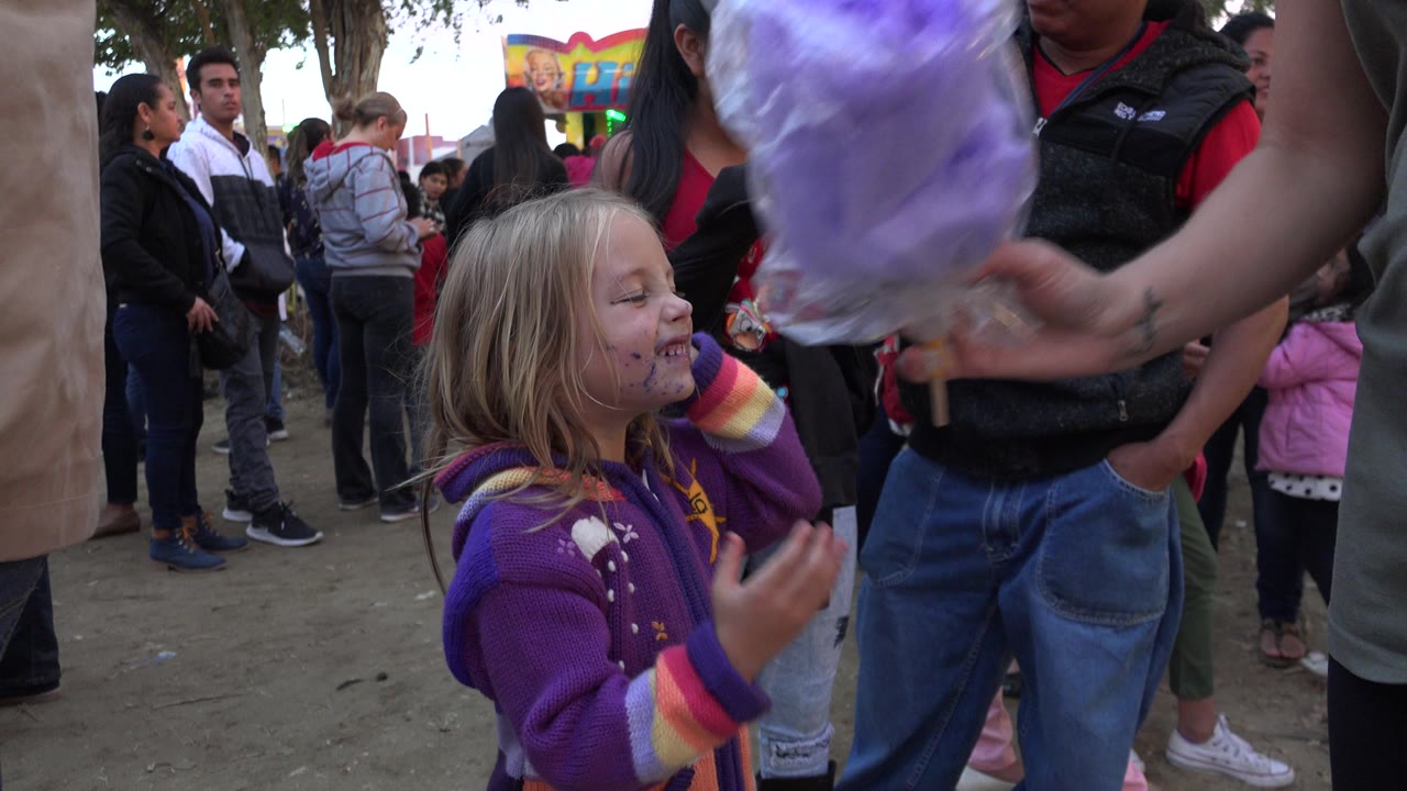 At 0:01 in San Jose Del Cabo, Mexico, the child smiles and raises a hand while a purple cotton candy bag partly covers the upper frame and a crowd fills the background.