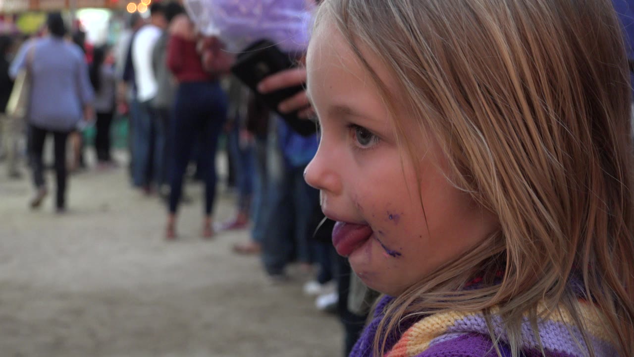 Young girl in profile licking purple cotton candy in a crowd.