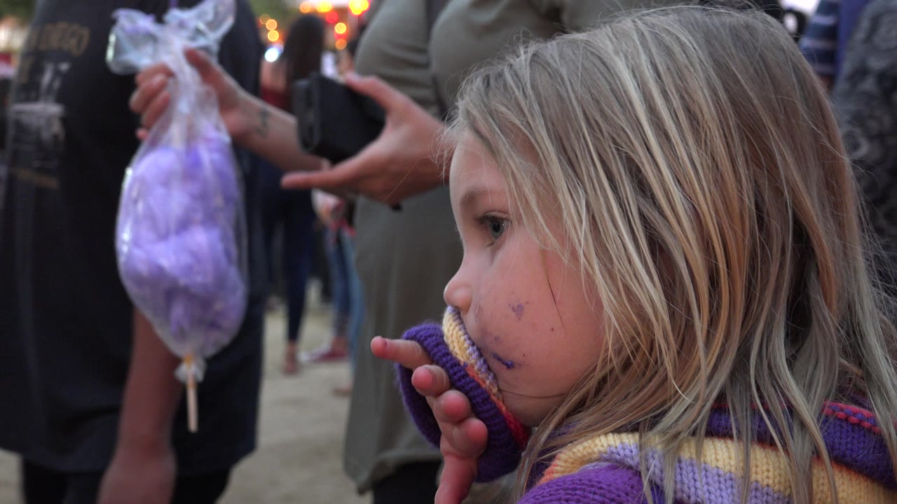 Blonde child in a striped sweater wipes her mouth near adults holding cotton candy.