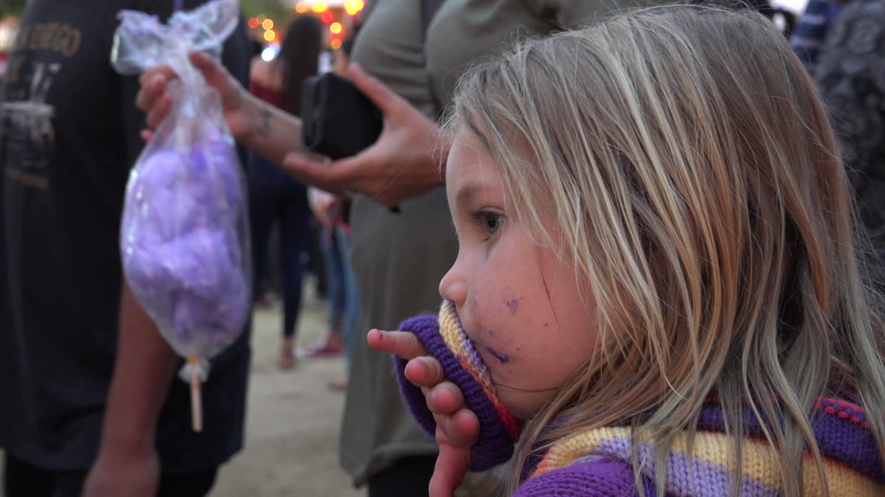 At 0:01 in San Jose del Cabo, Mexico, the child remains in profile with her fingers near her mouth while a plastic bag of purple cotton candy hangs behind her.