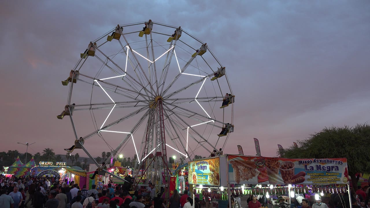 At 0:02, the white-lit Ferris wheel is framed slightly right of center with fair booths, crowd movement, and colorful signs below in San Jose del Cabo.