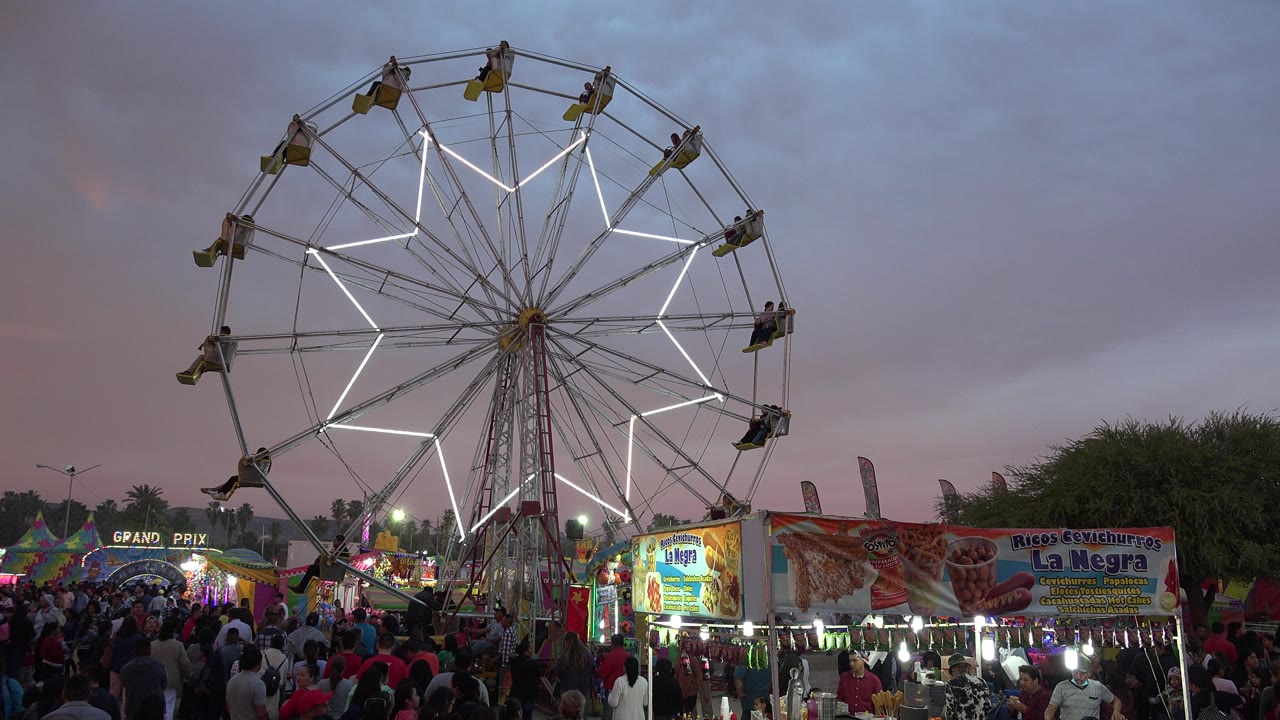 Lit Ferris wheel above a crowded carnival at dusk.