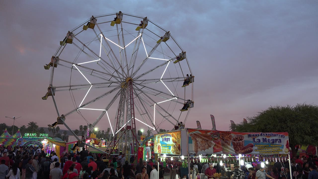 A lit Ferris wheel towers over a crowded fairground in San Jose del Cabo, Mexico, with vendor stalls and signage below a darkening sky.