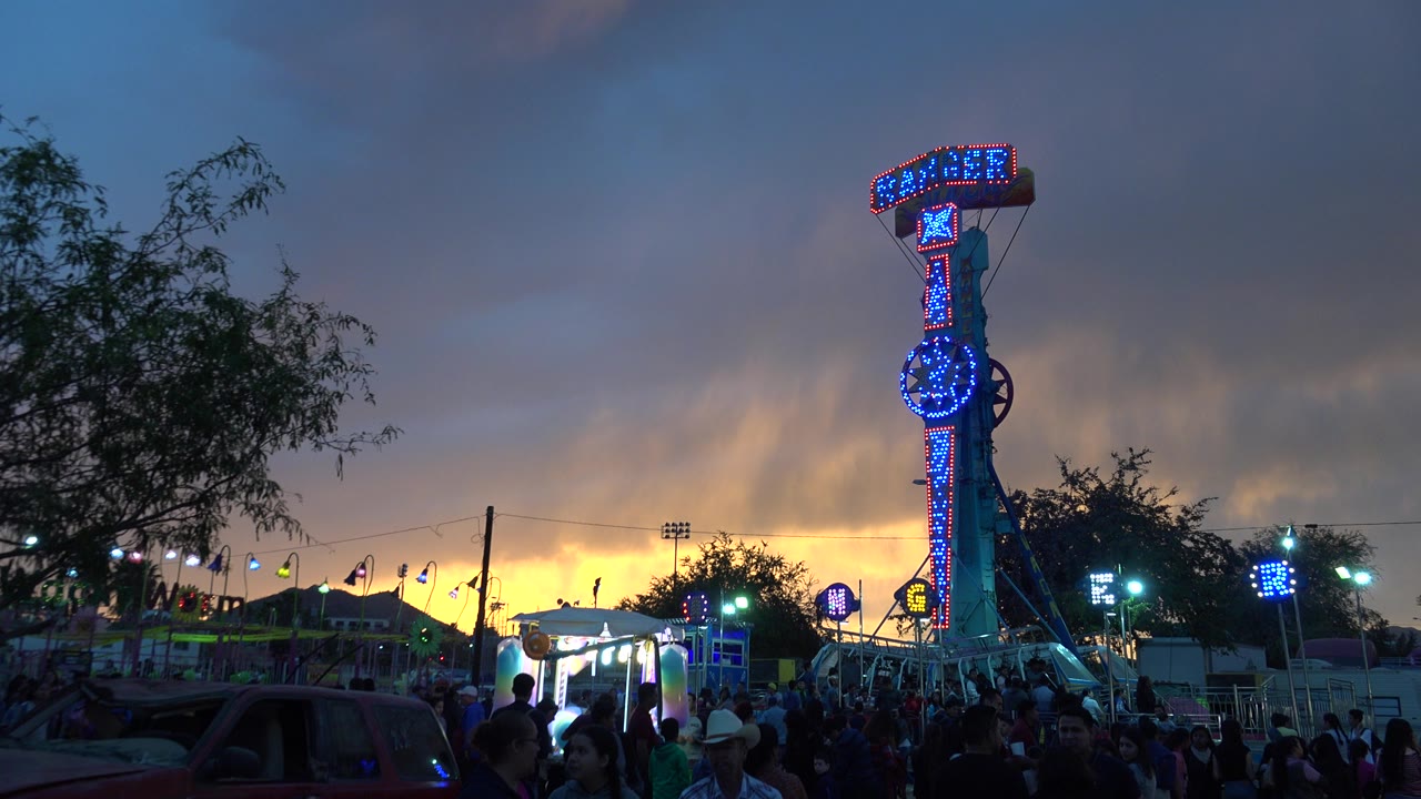 In San Jose del Cabo, Mexico, an illuminated blue ride tower stands above a crowd, tents, trees, and a glowing orange dusk horizon.