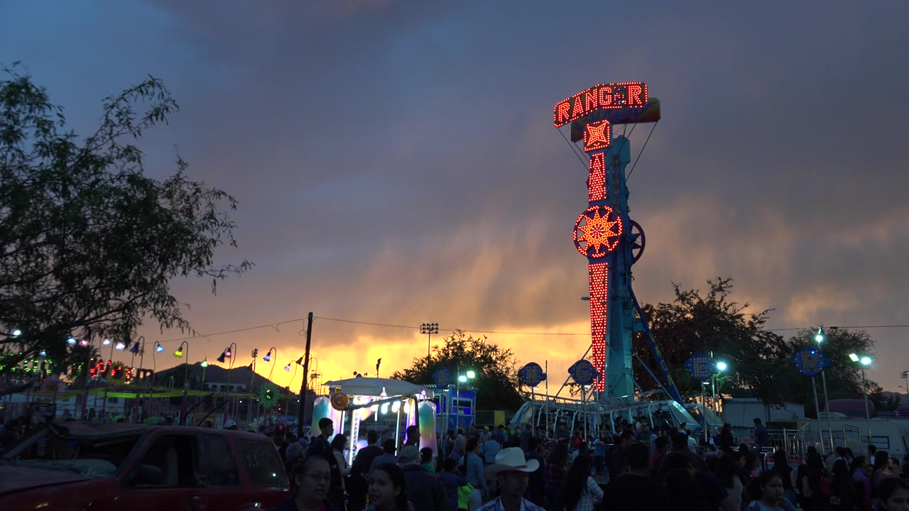 The San Jose del Cabo fairground remains busy with people in the foreground, ride lights across the midway, and the Ranger sign standing against a bright sunset band.