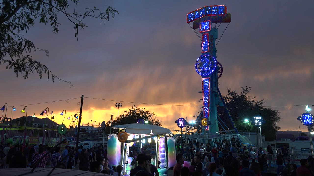 At 0:00, a blue-lit Ranger ride sign stands over a San Jose del Cabo fairground with a bright sunset band, tree branches, stalls, and distant people visible below.