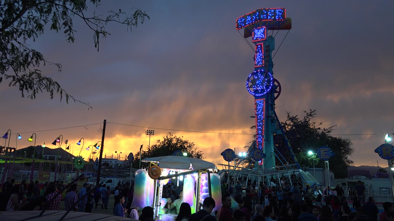 The ride remains lit in blue and red while people gather near bright foreground booths and the darkening San Jose del Cabo sky fills the background.