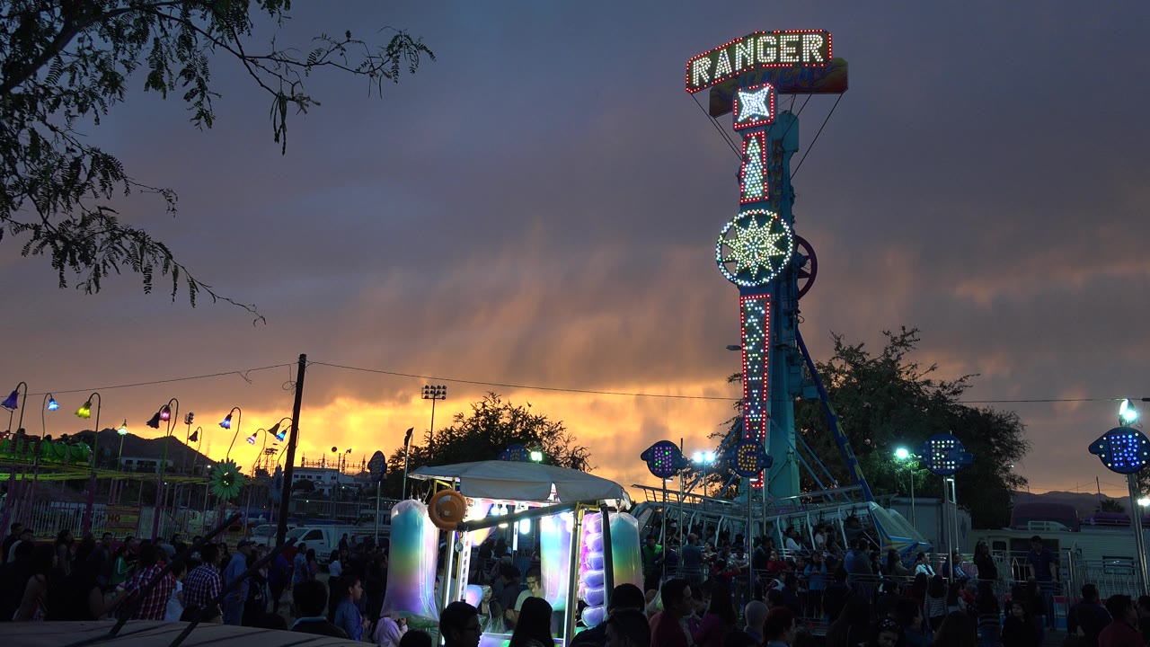 The Ranger ride stands near center with bright red and green lights, fair booths and visitors below, and a warm sunset strip along the horizon in San Jose del Cabo, Mexico.