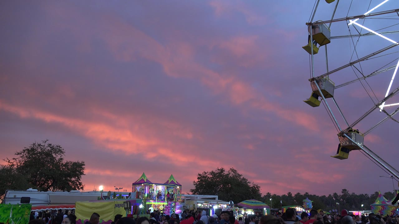 A San Jose del Cabo carnival crowd gathers beneath pink sunset clouds, with booths in the midground and a cropped ferris wheel structure at the right edge.