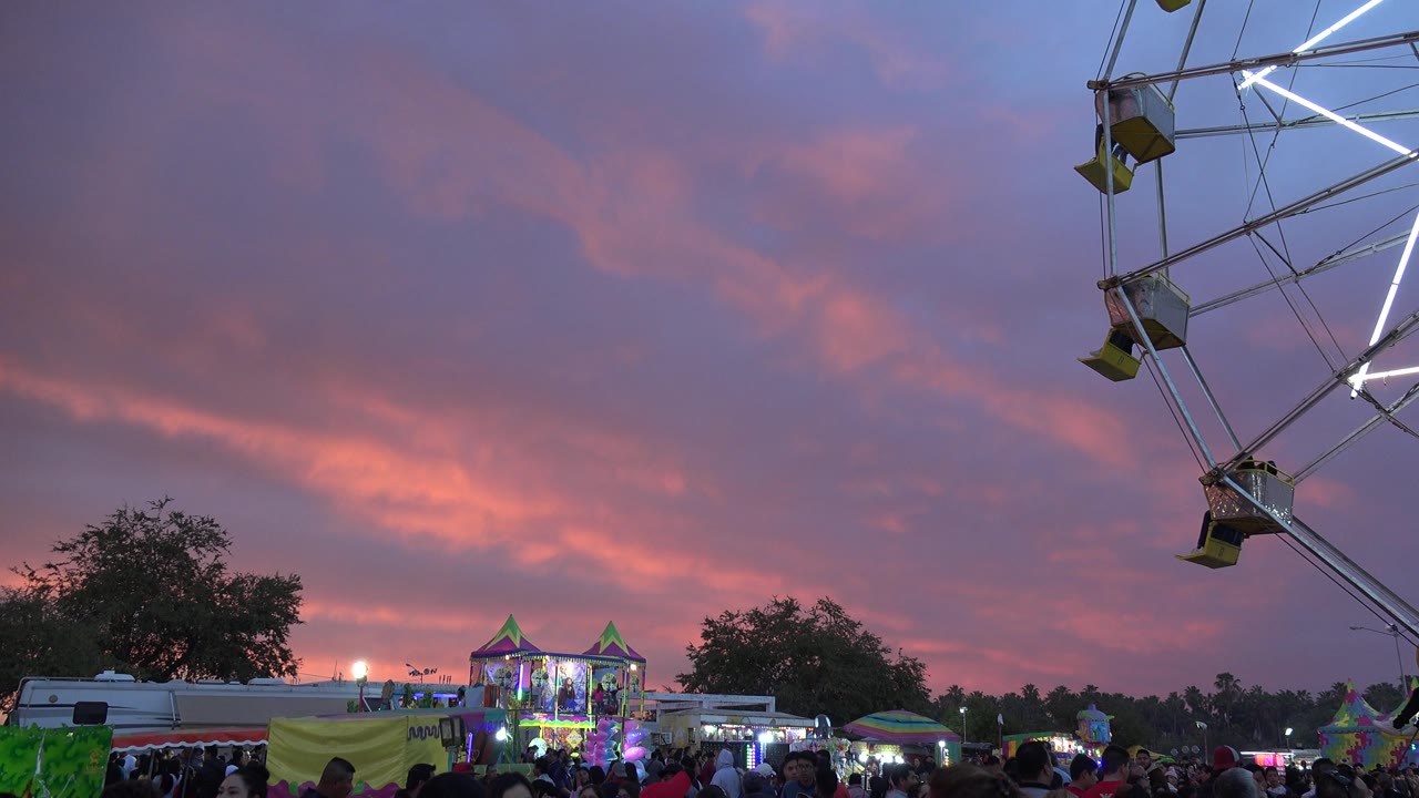 The San Jose del Cabo fairground remains busy at 0:01, with people along the bottom of frame, lit carnival booths near center, and the ferris wheel cropped on the right.