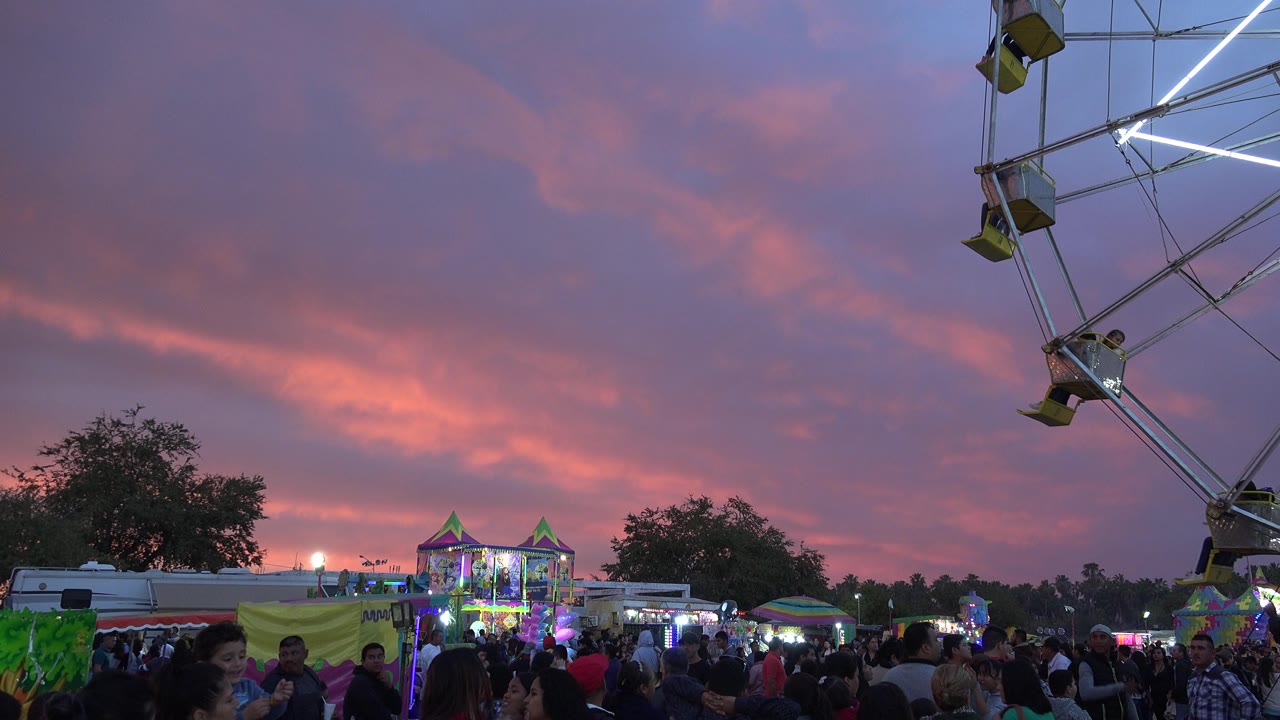 Crowd at dusk with Ferris wheel frame and pink clouds overhead.
