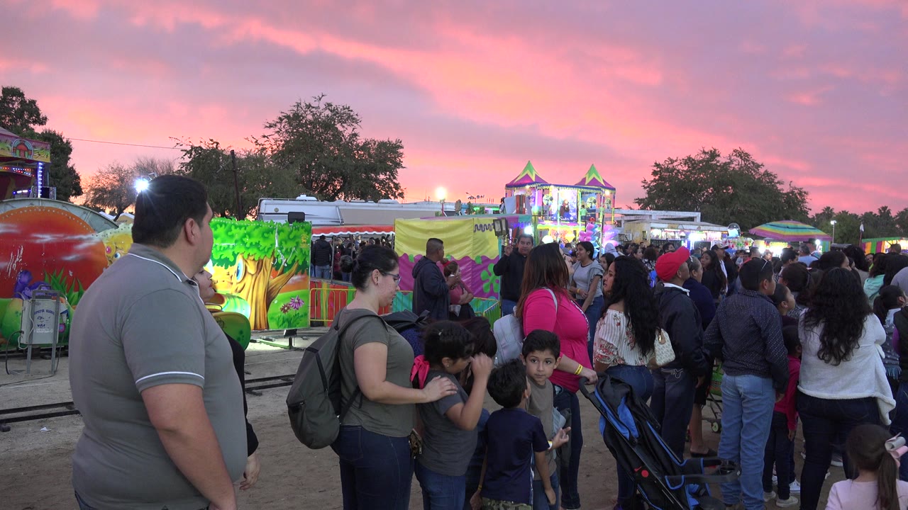 Families standing in a carnival line under a pink dusk sky.