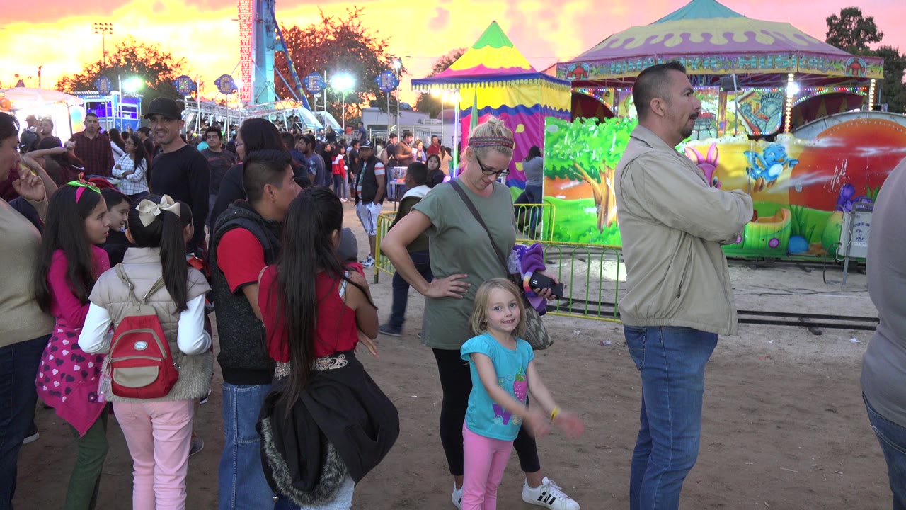 A group of children and adults fills the foreground in San Jose del Cabo, Mexico, while a blonde child faces the camera near a man in a beige jacket and a painted ride car.