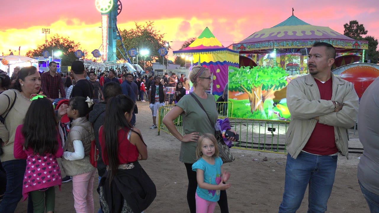 Adults and children wait in a ride queue in San Jose del Cabo, Mexico, with a painted carnival facade, bright ride lights, and a pink orange sunset sky behind them.