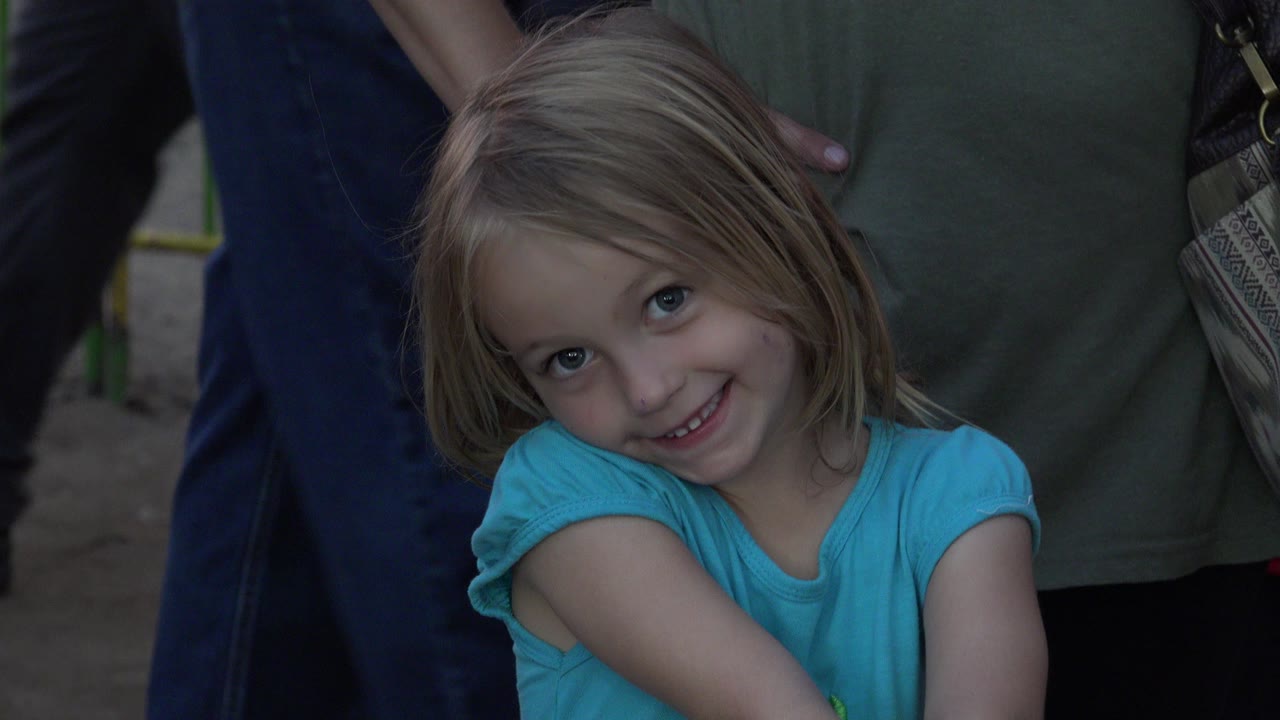 A blonde young girl in a blue shirt smiles toward the camera in San Jose del Cabo, Mexico, with adult torsos and a fenced dirt area behind her.