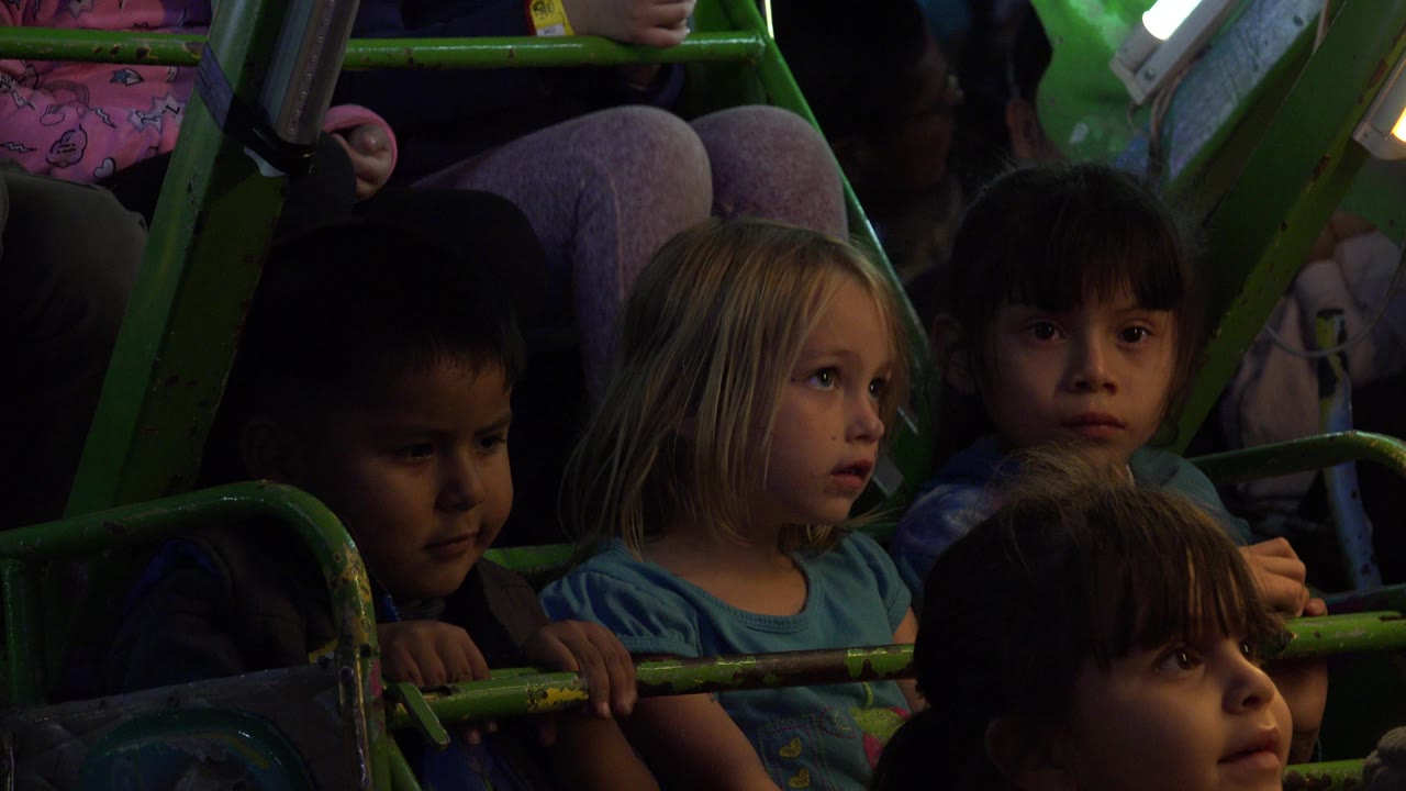 Children seated behind green amusement ride bars in dim light.