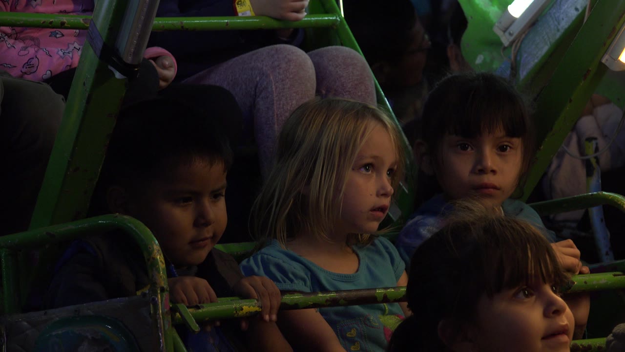 A group of children remain seated behind the green ride rail in San Jose Del Cabo, Mexico, with the center girl looking ahead and a child in front partly blocking the lower frame.