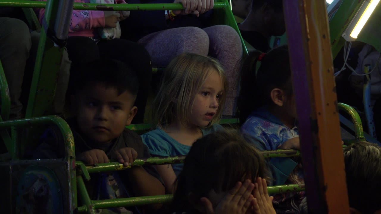 Children seated behind green carnival ride bars in dim light.