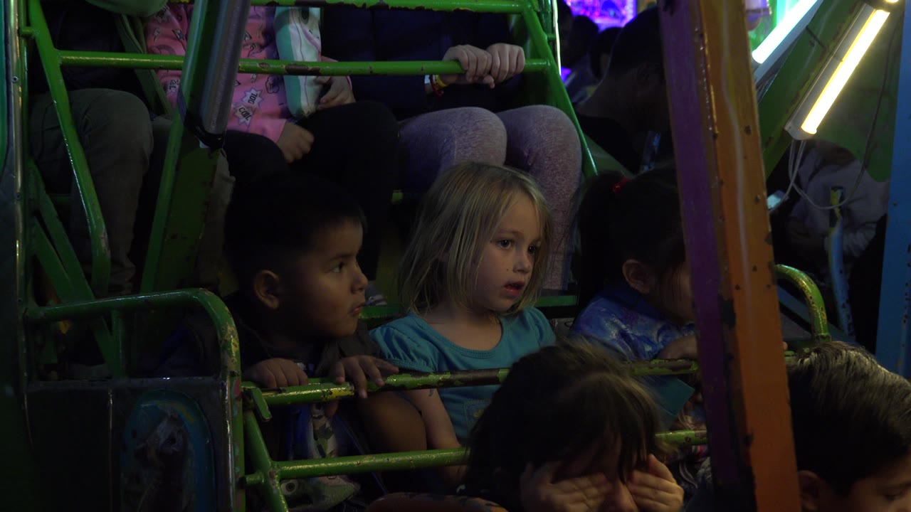The children remain seated in the carnival ride in San Jose Del Cabo, Mexico, with one child looking sideways and bright ride lighting cutting across the upper frame.