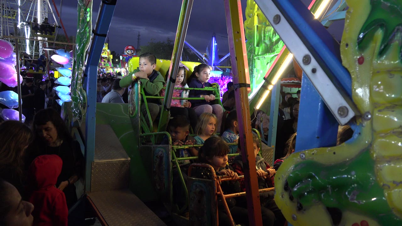 A group of children sits inside the green ride structure in San Jose del Cabo, Mexico, while adults and bright carnival lights fill the background.