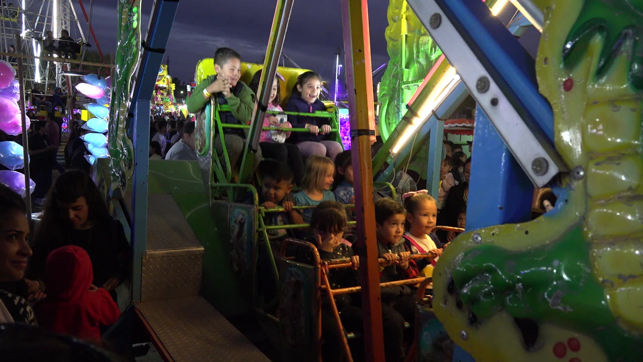 Children seated on a green amusement ride at night with adults nearby.