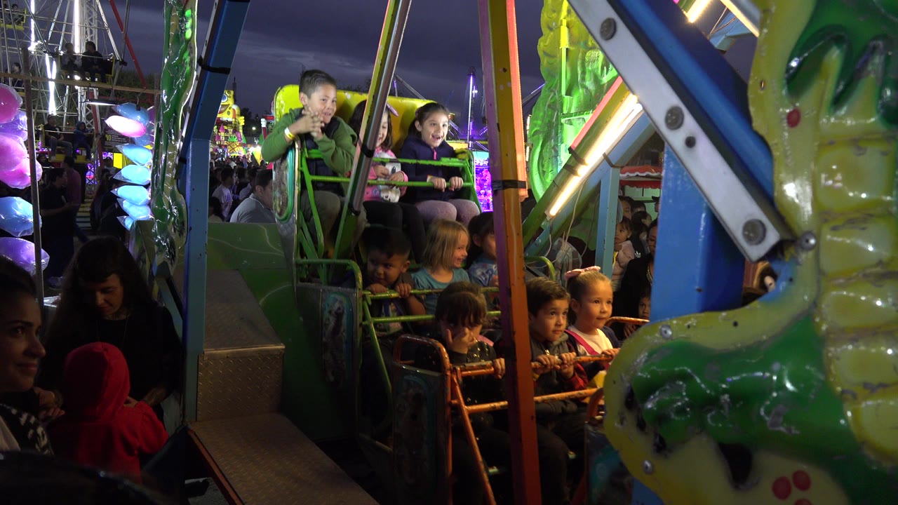 Children on the San Jose del Cabo amusement ride are framed by green railings and a large animal-shaped ride panel, with crowded night lights in the background.