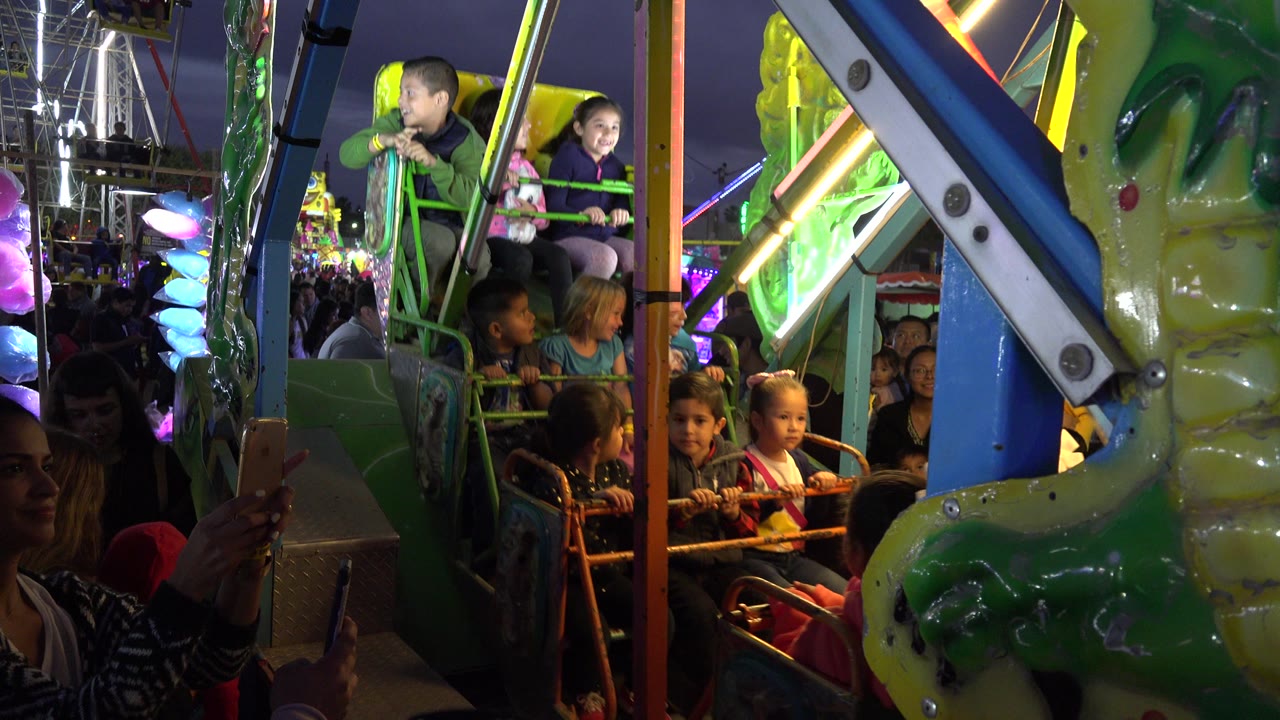 Children ride a colorful amusement ride at night with spectators nearby.