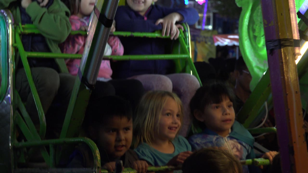 Three children remain centered behind the green ride barrier as a bright green panel and dim background lights frame the amusement park setting in San Jose Del Cabo, Mexico.