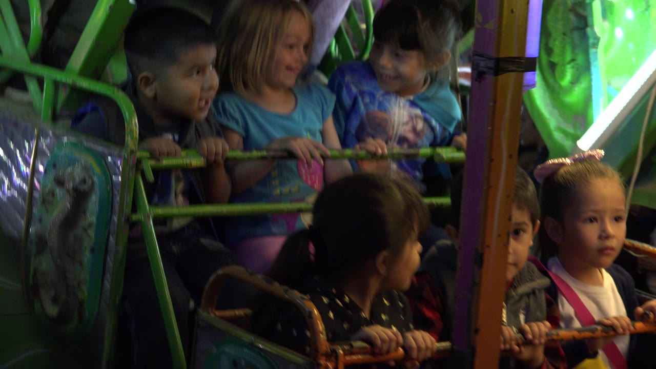 Several children hold the green safety bar on the carnival ride in San Jose del Cabo, Mexico, while bright white and green light fixtures glow behind them.